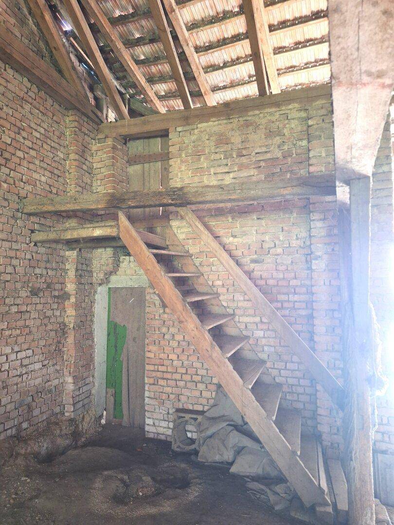 Rustikaler Innenraum einer Scheune mit Ziegelwänden, Holzbalken und einer steilen Holztreppe zum Obergeschoss.