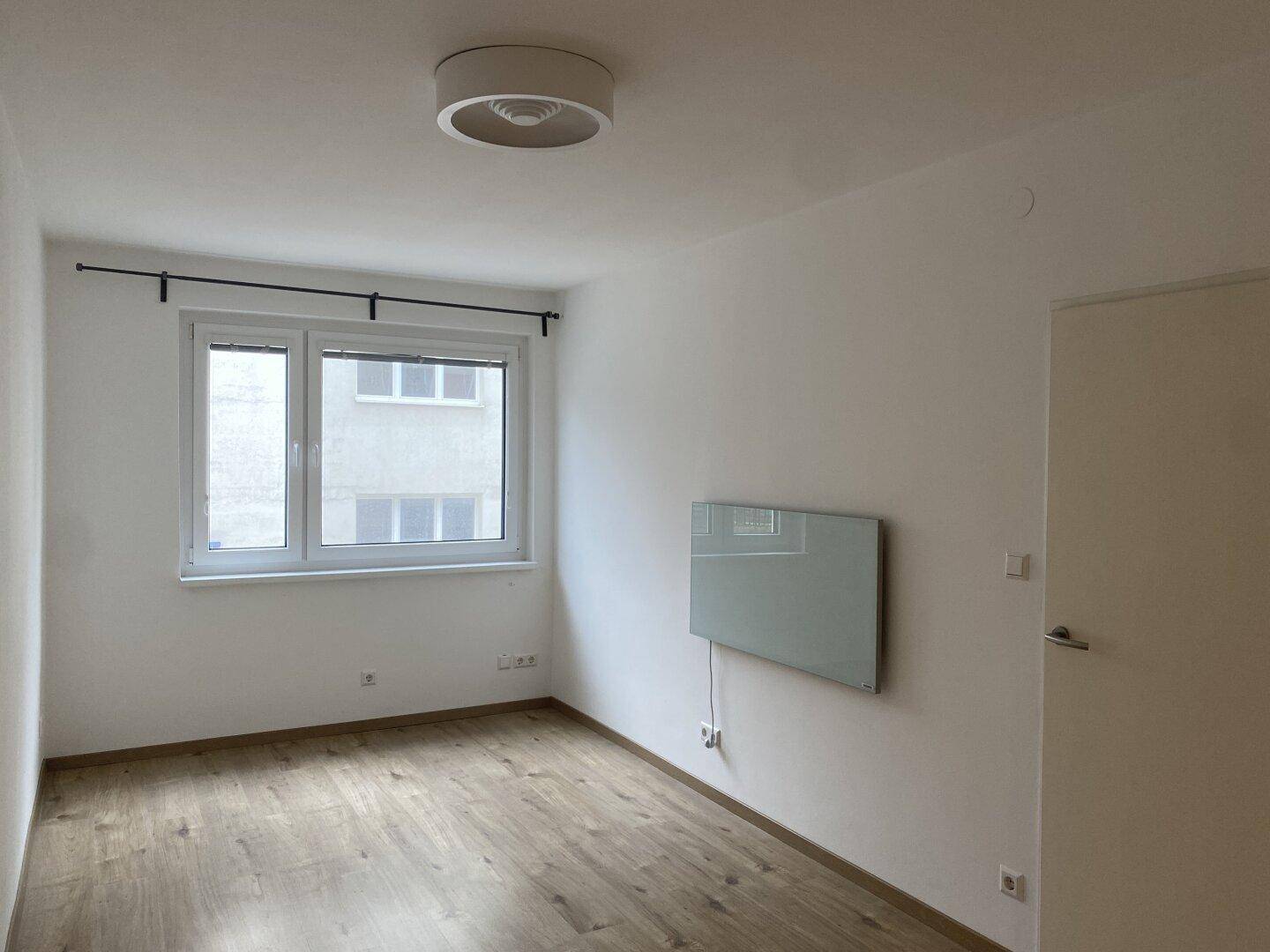 Leeres Schlafzimmer mit Holzfußboden, großem Fenster und moderner Wandheizung.