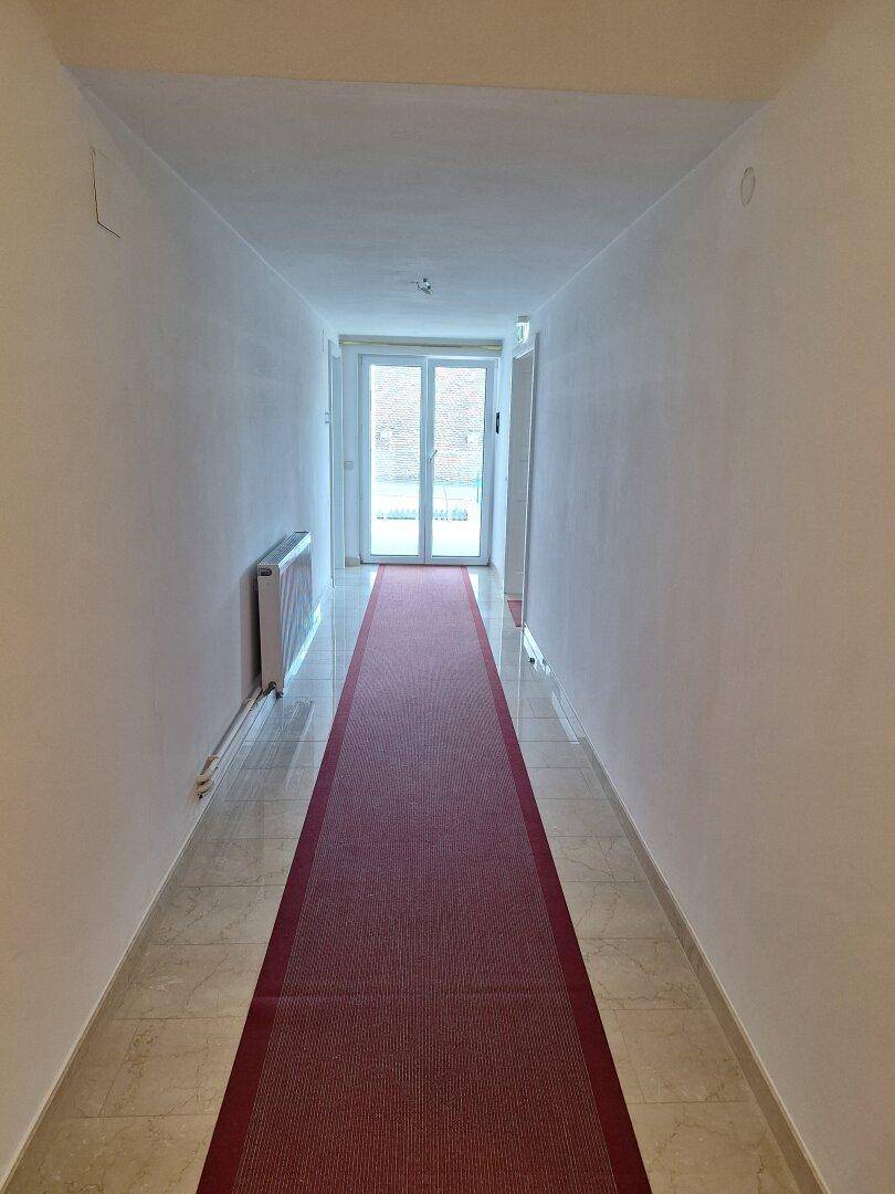Langer, heller Flur mit Fliesenboden, rotem Läufer und Zugang zu einem Außenbereich.