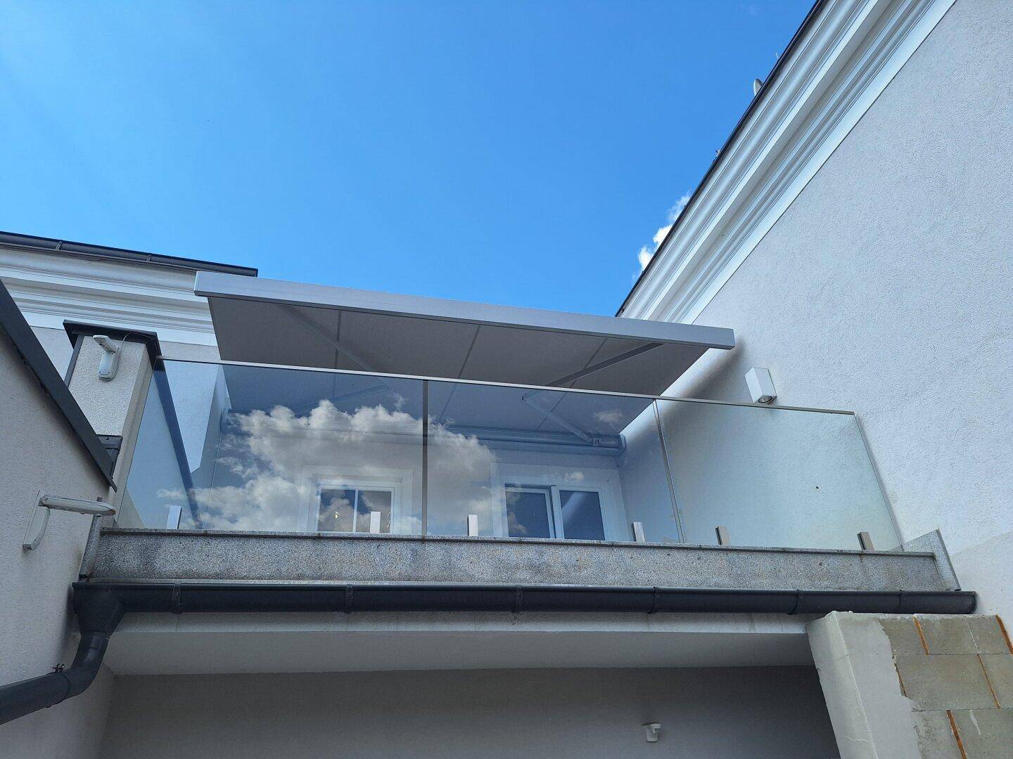 Moderner Balkon mit Glasgeländer und ausfahrbarer Markise unter blauem Himmel.