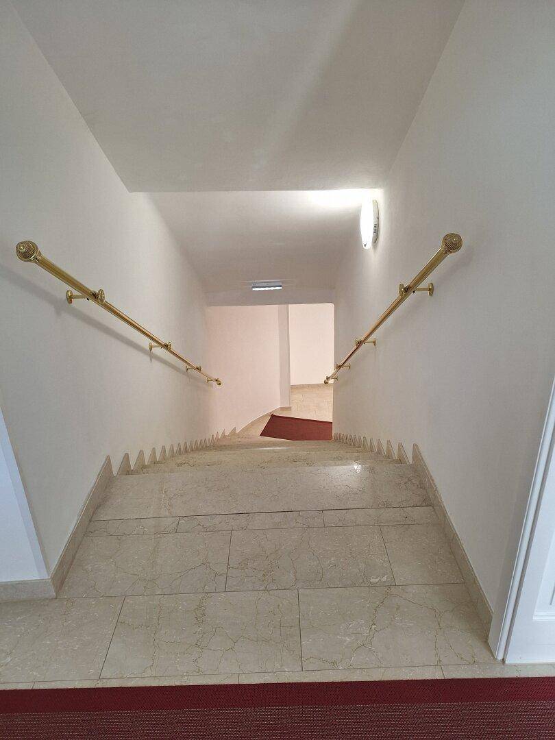 Heller Treppenabgang mit Fliesenstufen, goldenen Handläufen und einem roten Teppichläufer.