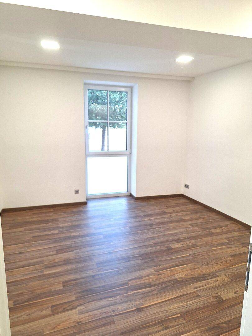 Leerer Raum mit Holzboden und einem großen Fenster, das viel Tageslicht hereinlässt.
