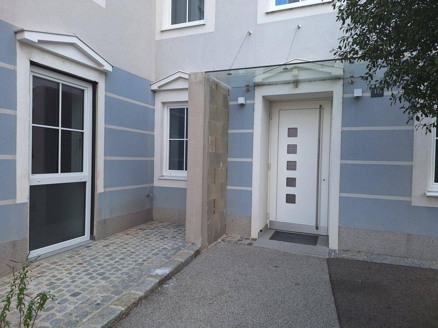 Eingangsbereich eines Gebäudes mit blauer Fassade, weißer Tür und einer Rampe für Barrierefreiheit.