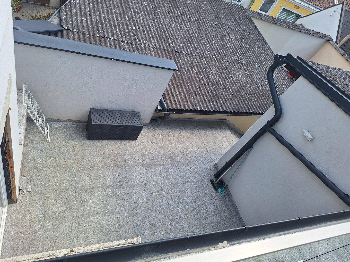 Blick von oben auf eine gepflasterte Terrasse mit einem Abstellkasten und angrenzenden Dächern.