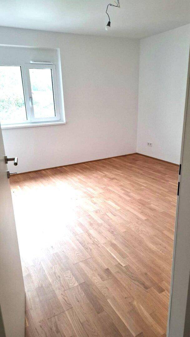 Leeres Schlafzimmer mit hellem Parkettboden und einem Fenster, das viel Tageslicht hereinlässt.