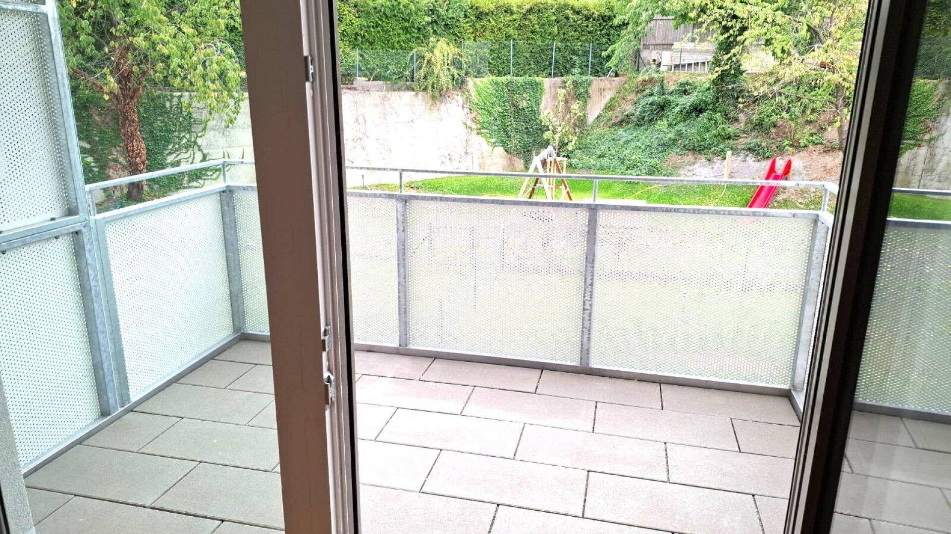 Heller Balkon mit Fliesenboden und Blick auf einen grünen Gartenbereich mit Spielgeräten.