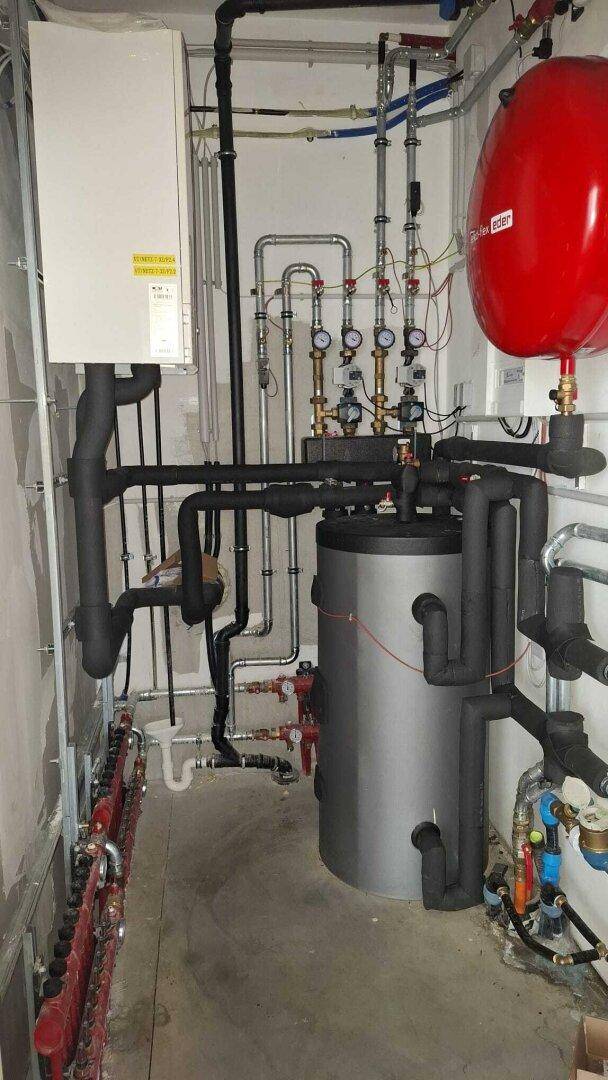 Technikraum mit zahlreichen Rohren, einem Warmwasserspeicher und weiteren Installationen für die Gebäudetechnik.