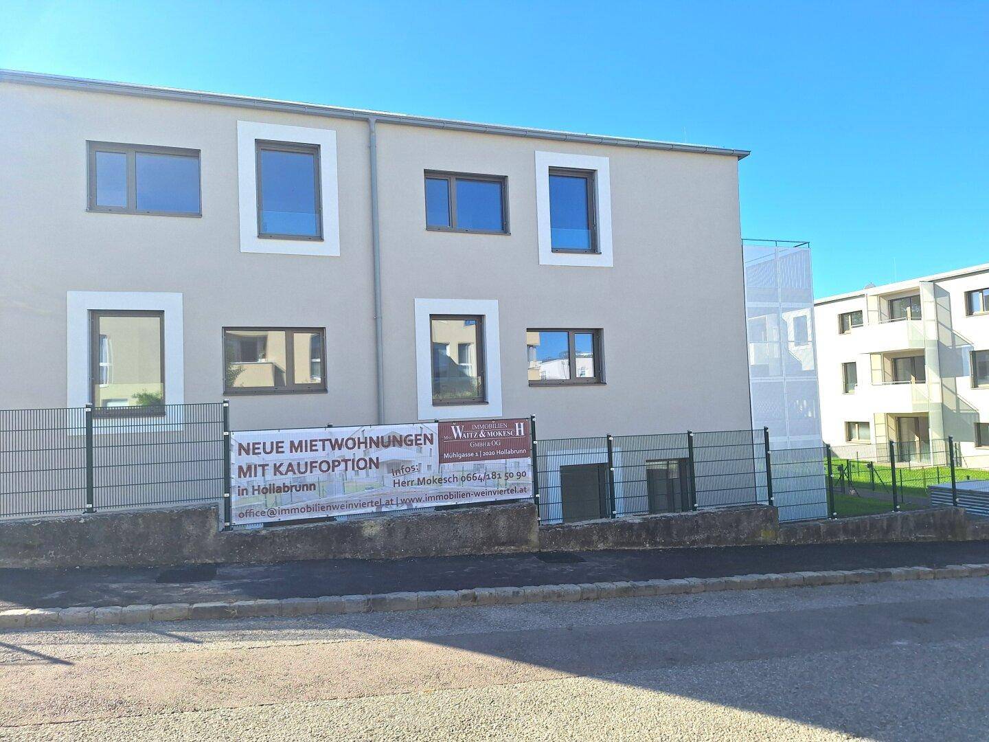 Ansicht eines modernen Wohngebäudes von der Straße mit Werbebanner im Vordergrund.