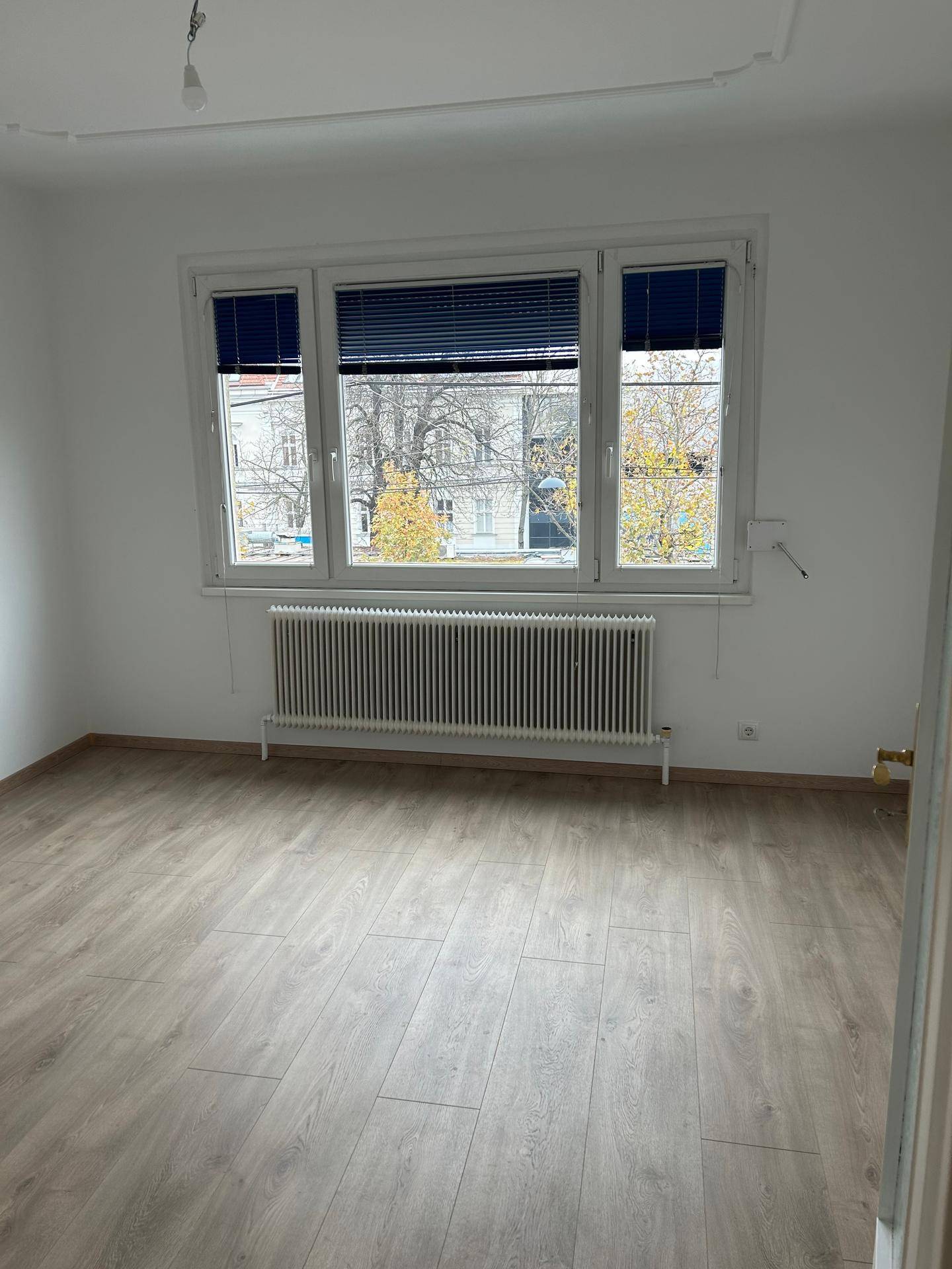 Geräumiges Zimmer mit Laminatboden, großen Fenstern und Heizkörper unter dem Fenster.
