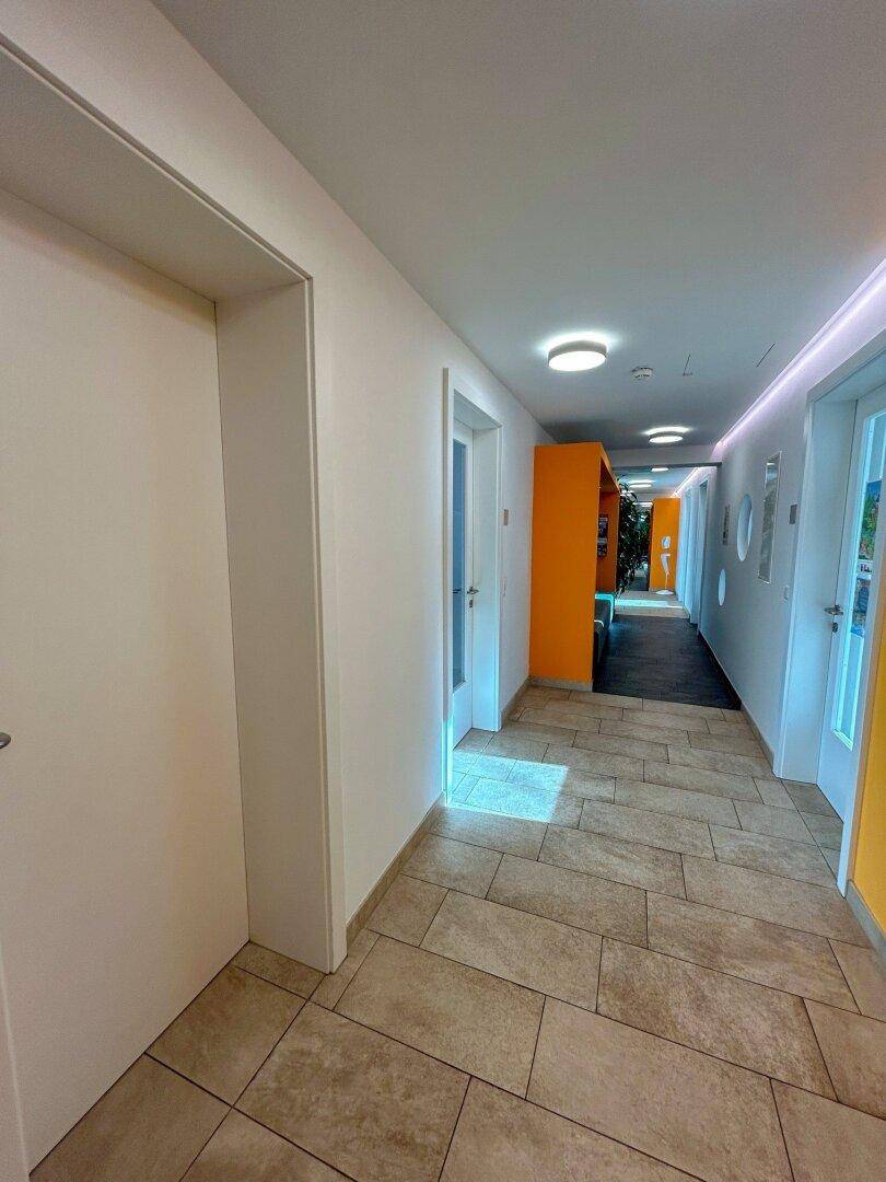Langer Flur mit hellen Fliesen, weißen Türen und orangefarbenen Akzenten im Hintergrund.