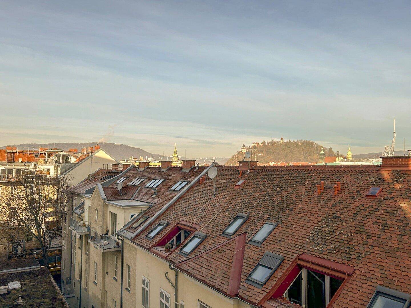 Blick über die Dächer der Stadt mit Schrägdachfenstern und Bergen im Hintergrund unter bewölktem Himmel.