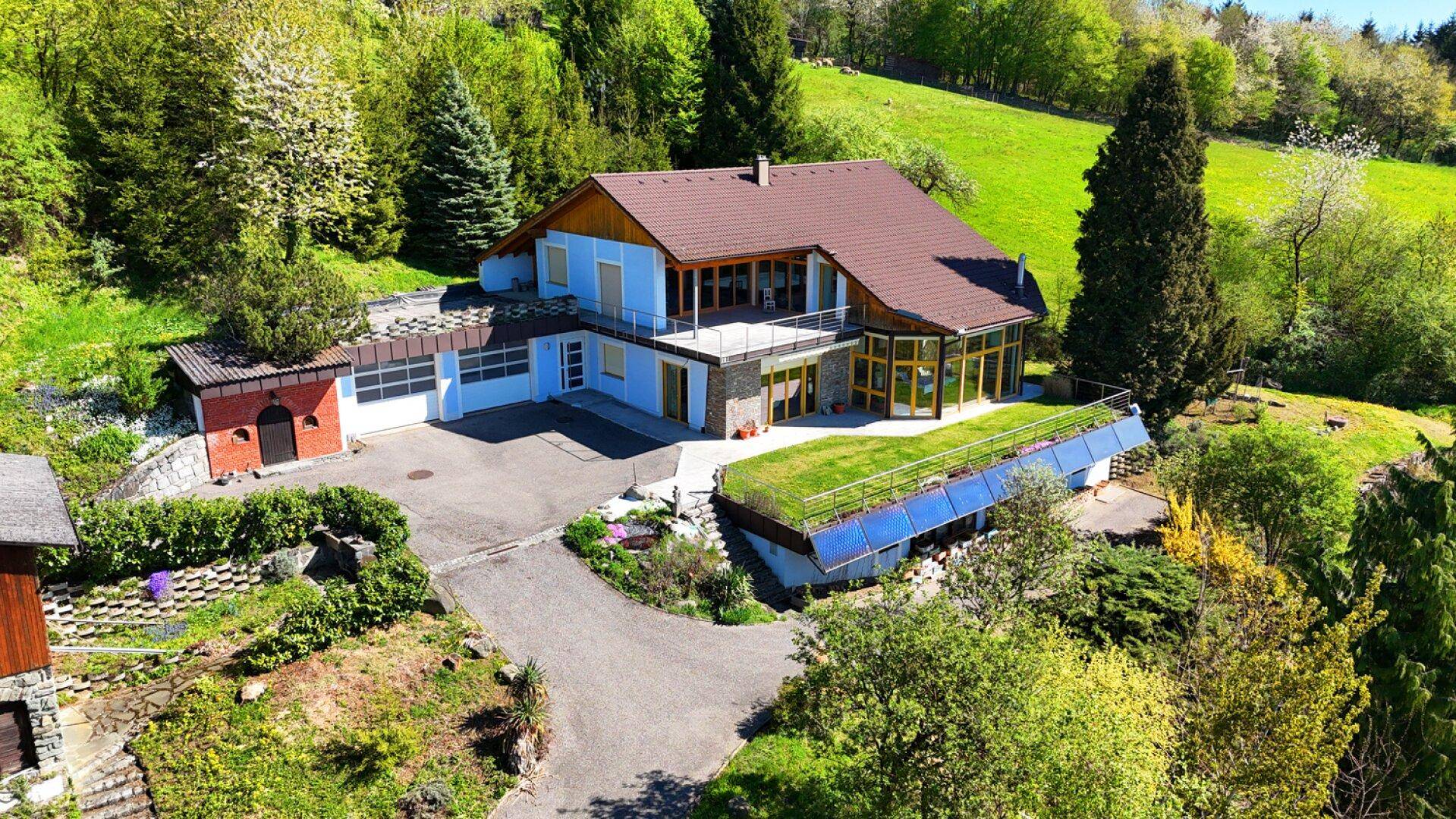 Luftaufnahme eines Hauses mit blauer Fassade, braunem Dach und umgebender grüner Landschaft.