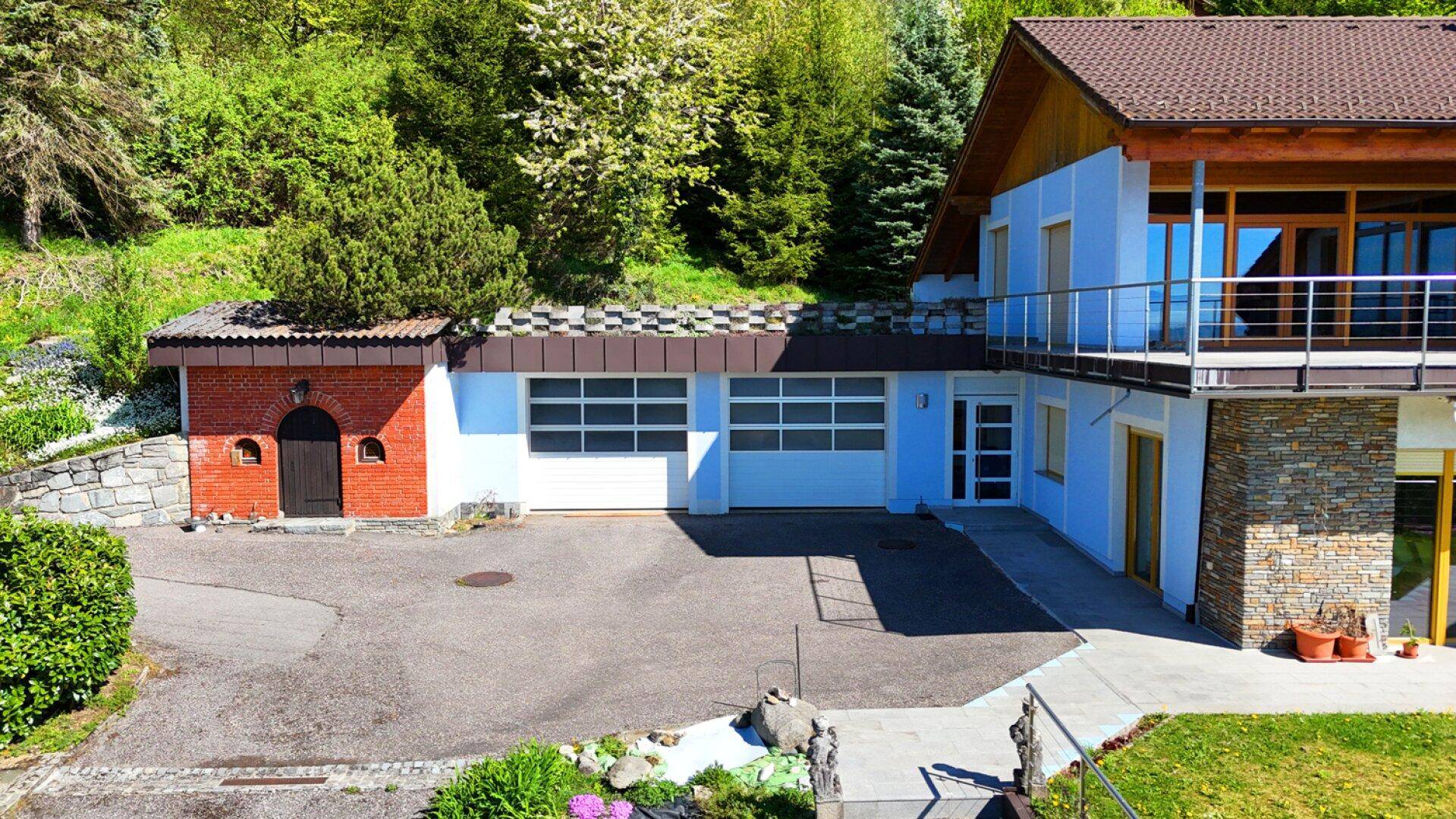 Ansicht der Doppelgarage und des Nebengebäudes mit roter Ziegelfassade und grüner Umgebung.