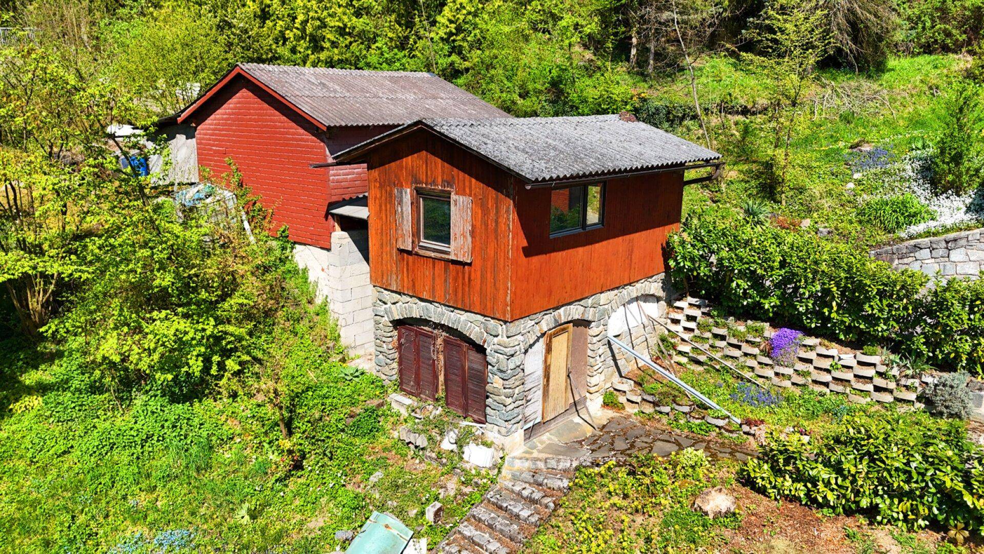 Rustikales Nebengebäude mit Holzwänden und Steinfundament, umgeben von viel Grün.