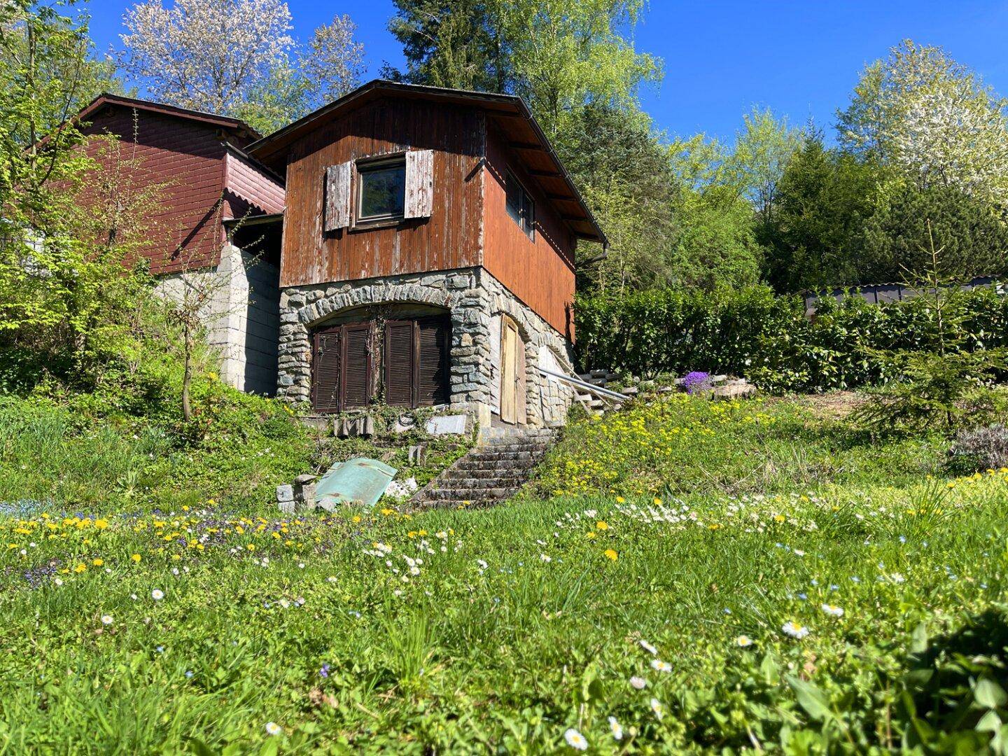 Rustikales Nebengebäude mit Holzwänden und Steinfundament, umgeben von einer Blumenwiese.