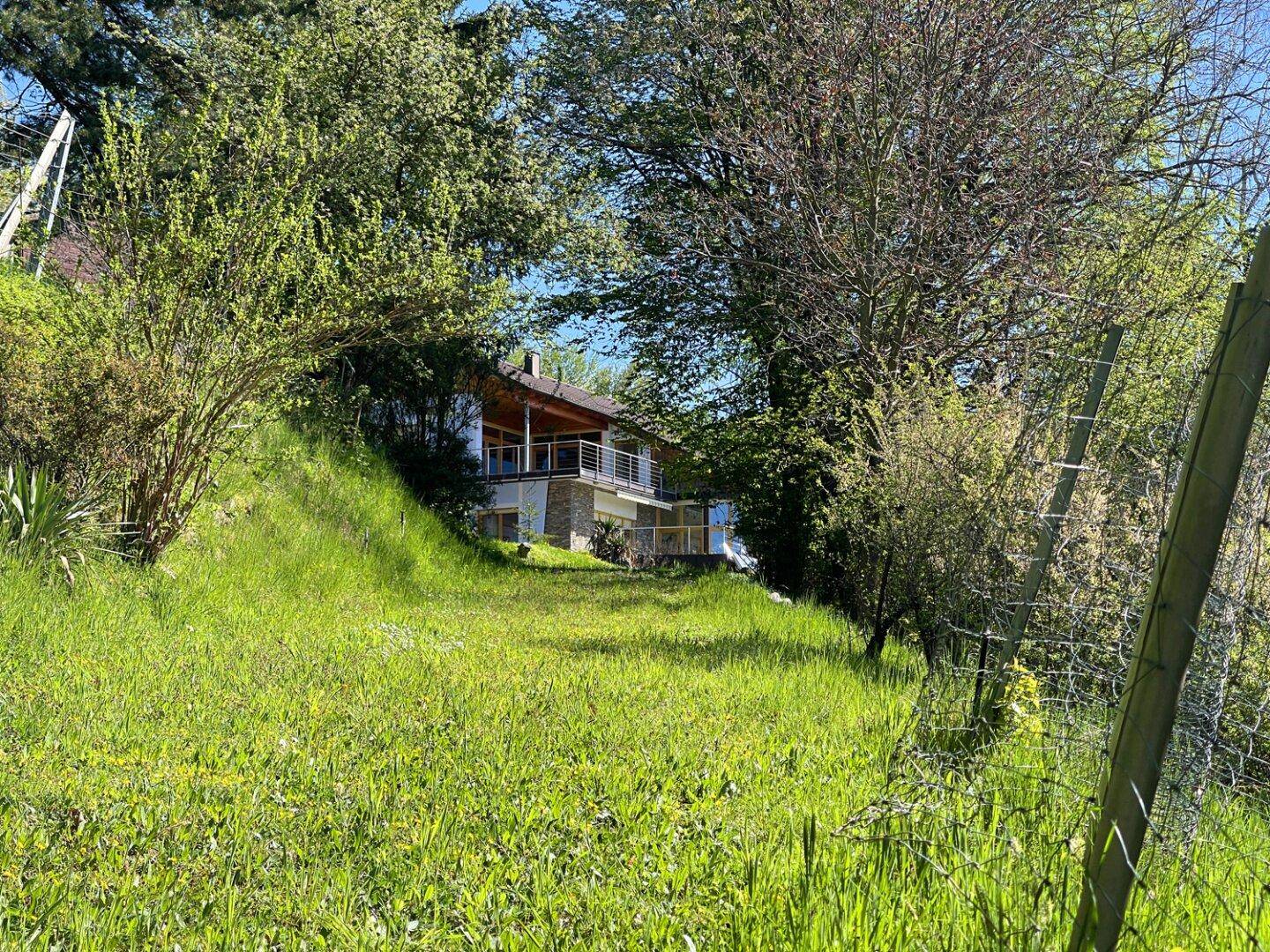Hausfassade mit Balkonen, umgeben von üppigem Grün und Bäumen auf einem Hanggrundstück.