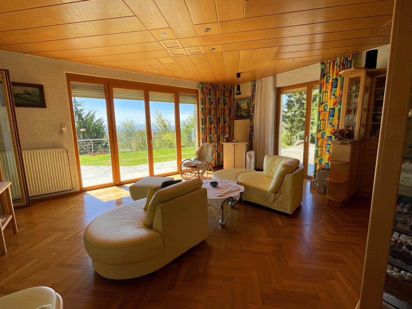 Lichtdurchflutetes Wohnzimmer mit Holzdecke, Fischgrätparkett und großen Fensterfronten mit Ausblick.