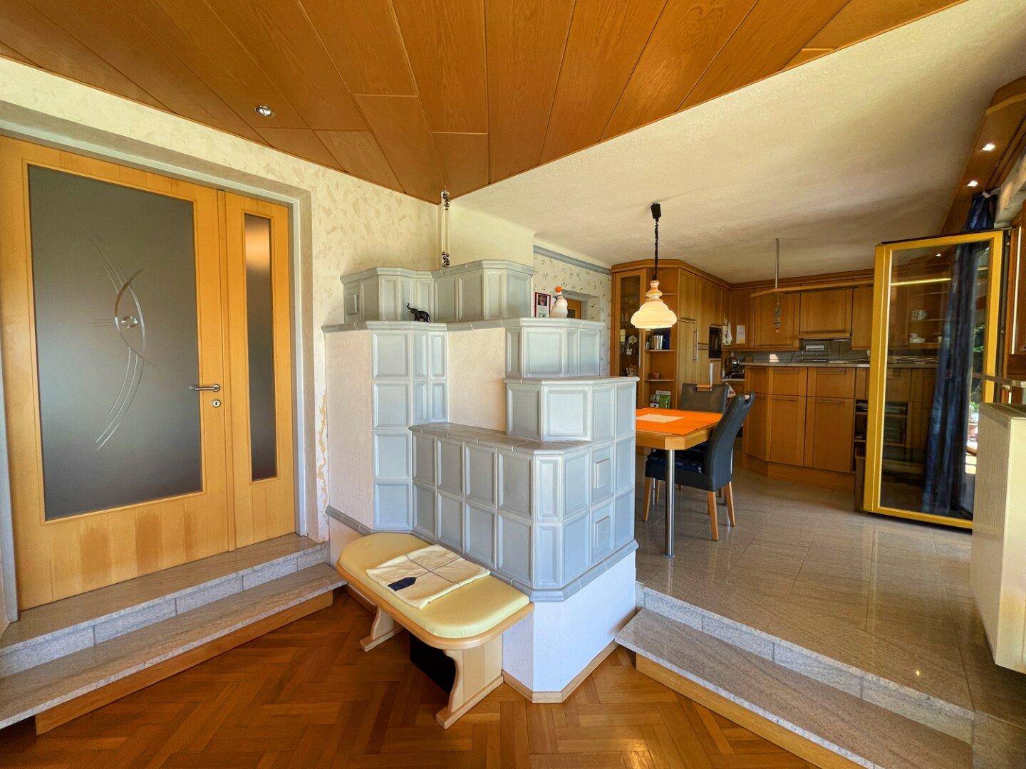 Übergang vom Wohnzimmer zur Küche, akzentuiert durch einen eleganten Kachelofen und Holzdetails.