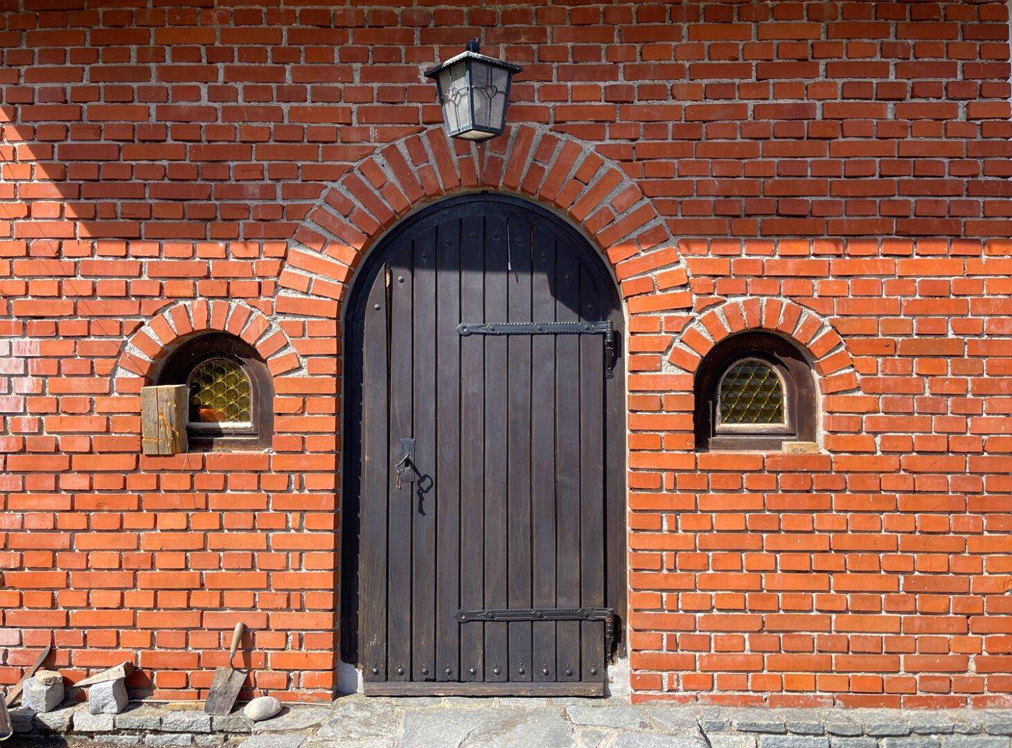 Rustikaler Eingangsbereich mit Holztür und Rundbogenfenstern in einer Ziegelmauer.