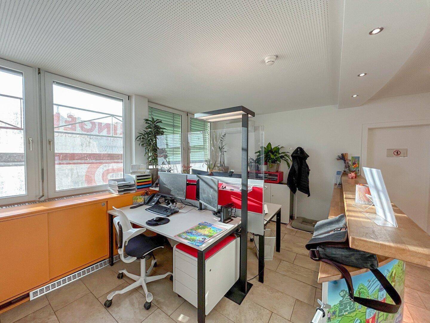 Moderner Bürobereich mit mehreren Arbeitsplätzen, großen Fenstern und orangefarbenen Akzenten.