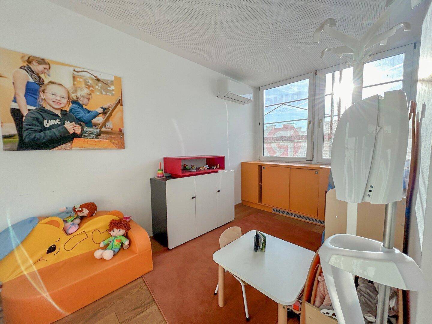 Freundlicher Spielraum mit Kindermöbeln, Teppichboden und großen Fenstern für viel Licht.