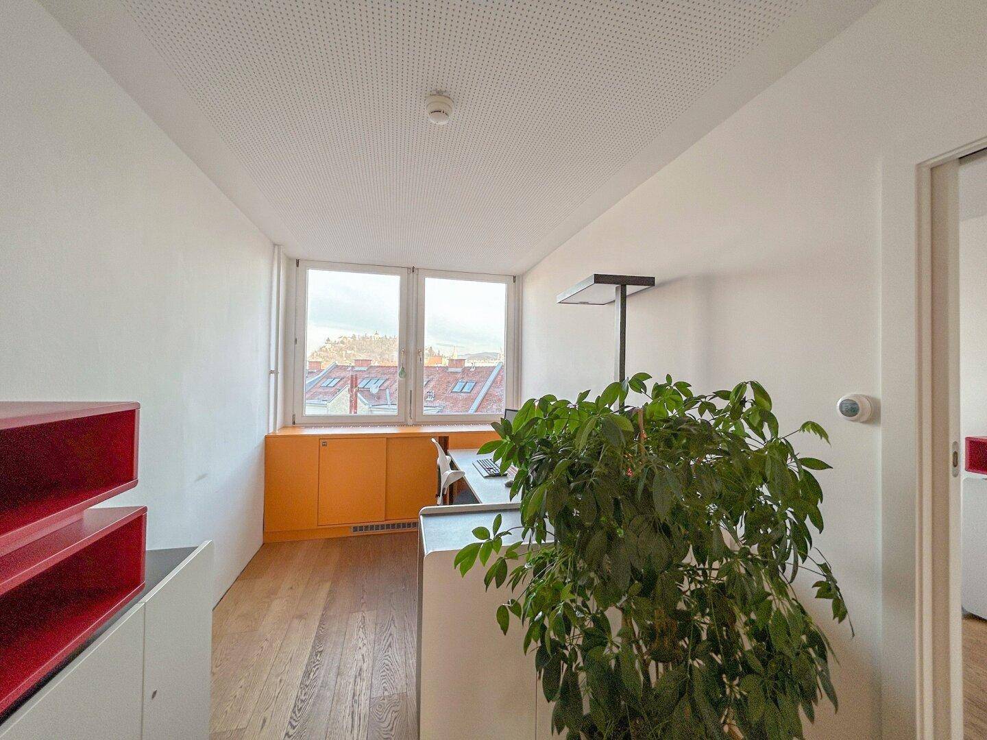 Helles Büro mit Holzboden, orangefarbenen Schränken und Blick auf die Dächer der Stadt.