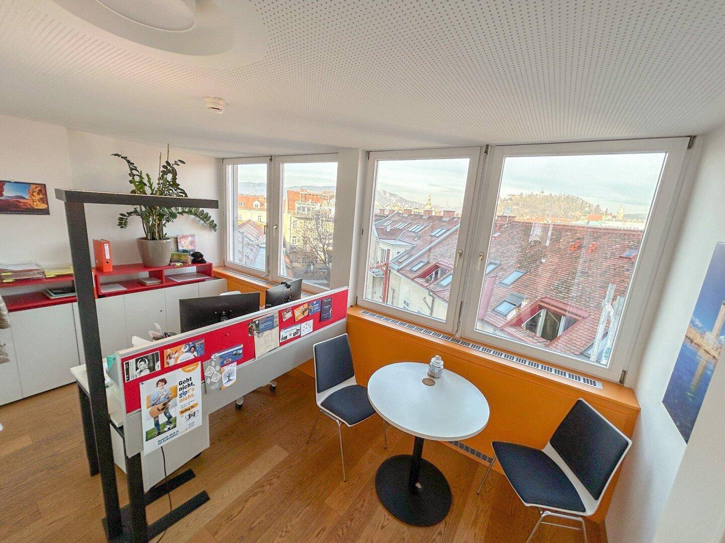 Heller Bürobereich mit Arbeitsplätzen, kleinem Besprechungstisch und weitem Blick über die Stadt.
