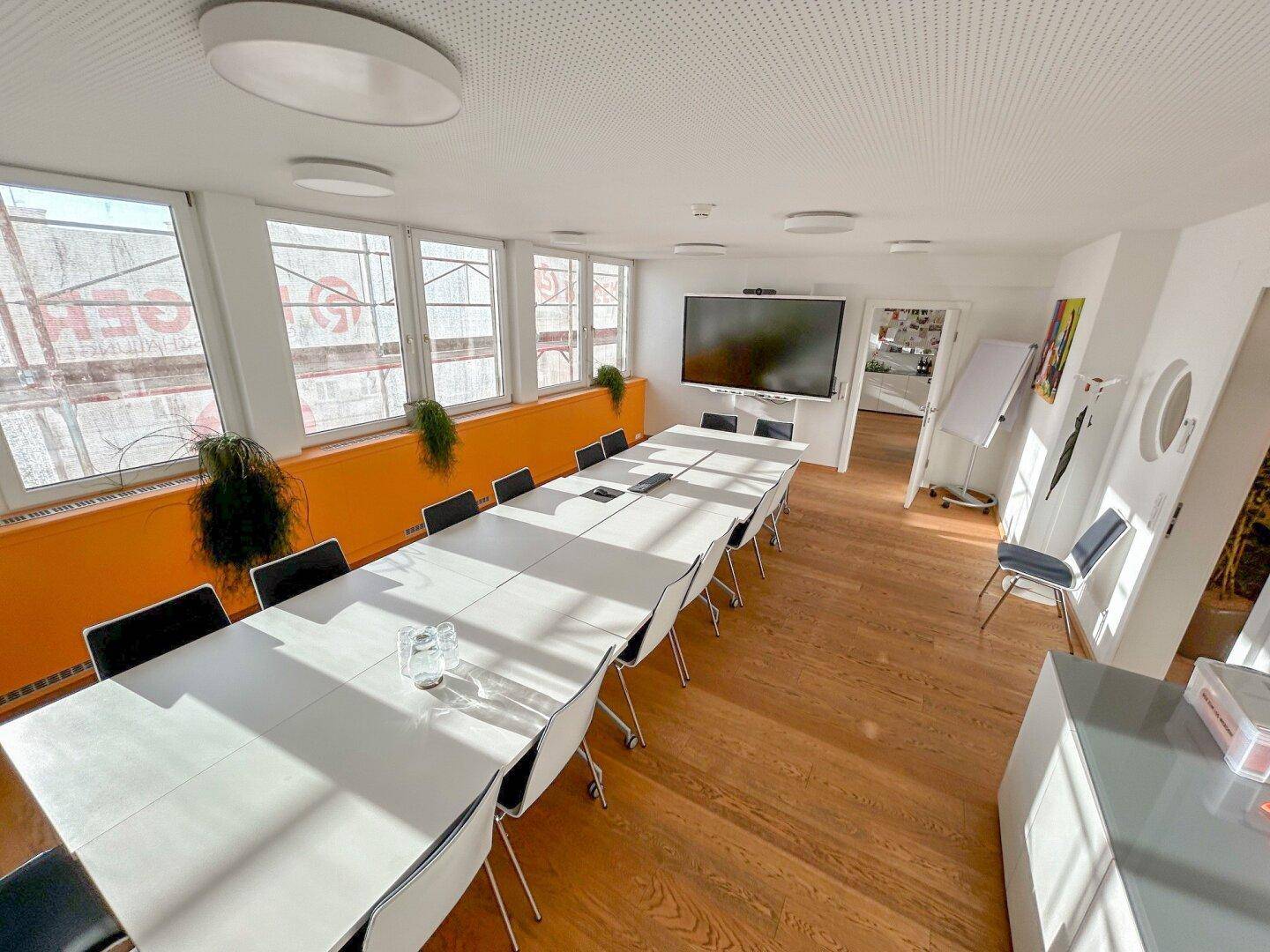 Geräumiger Konferenzraum mit langen Tischen, Stühlen, Holzboden und einem großen Wandbildschirm.