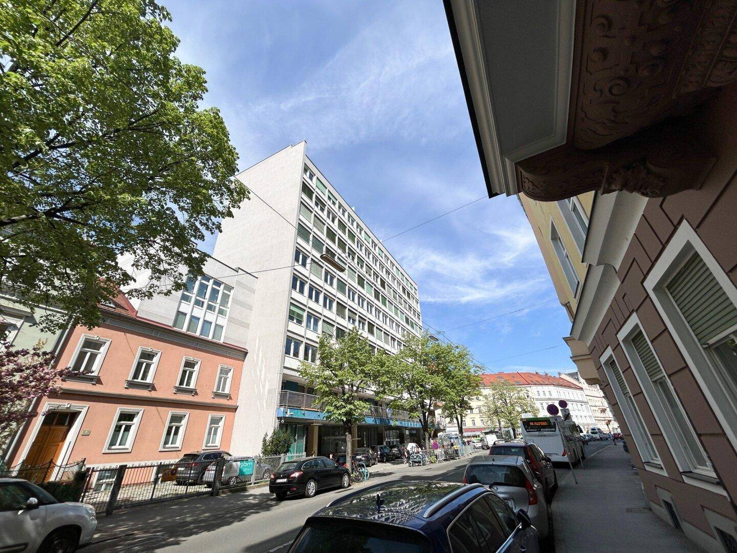 Modernes Gebäude an einer Straße mit Bäumen und geparkten Autos unter blauem Himmel.