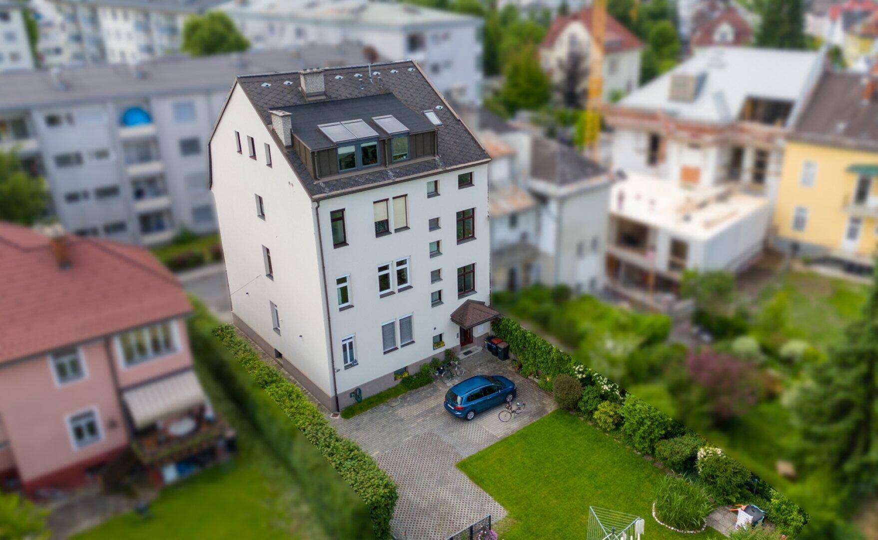 Schräge Luftaufnahme des Mehrfamilienhauses mit Garten, Einfahrt und umliegenden Gebäuden.