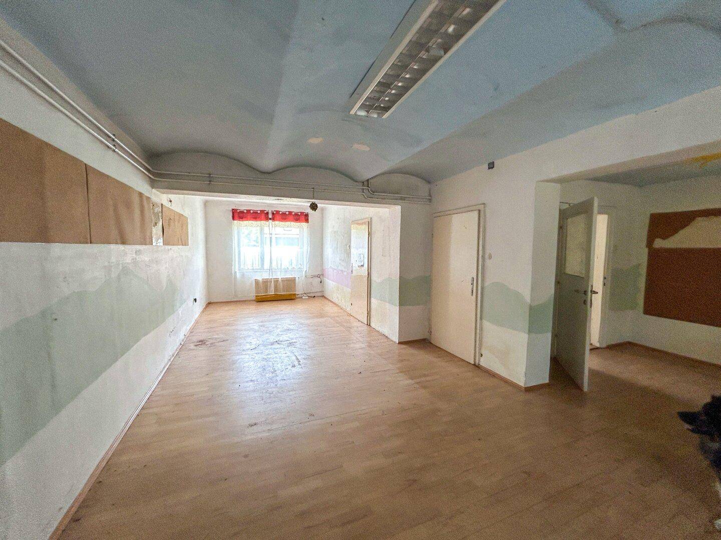 Großer, leerer Raum mit Holzboden und Fenstern, die viel Tageslicht hereinlassen.