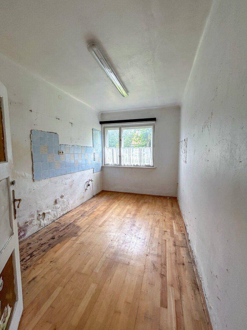 Leere Küche mit Holzboden, einem Fenster und Resten alter Fliesen an der Wand.