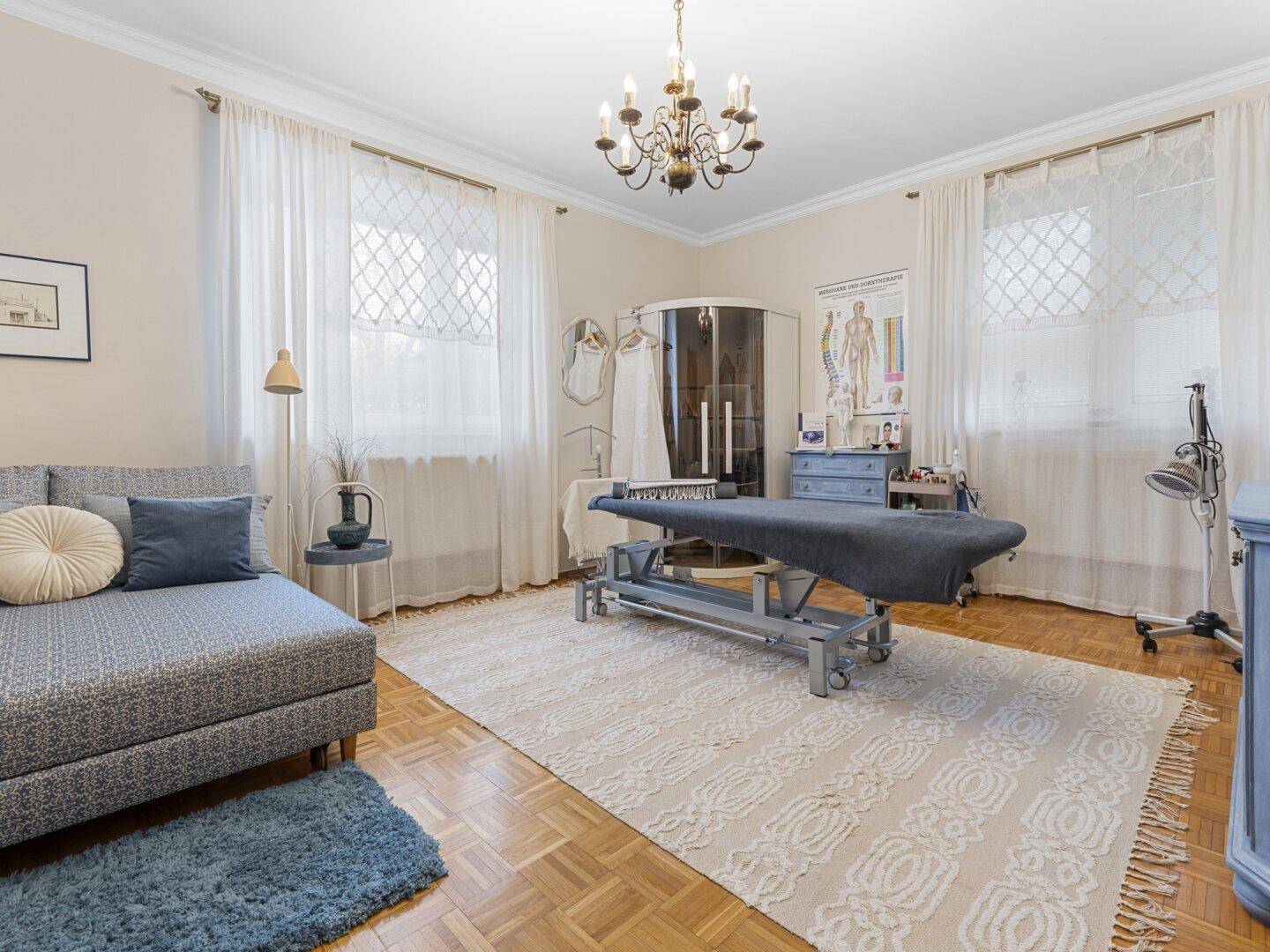 Geräumiges Zimmer mit Parkettboden, hohen Fenstern und einer Behandlungsliege, viel Tageslicht.
