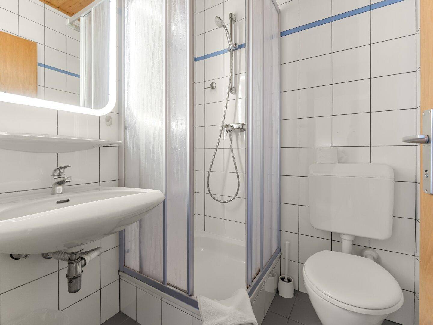 Helles Badezimmer mit weißen Fliesen, Waschbecken, Toilette und Duschkabine.