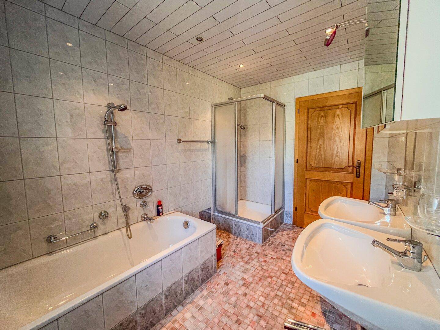 Badezimmer mit Badewanne, Duschkabine und Doppelwaschbecken, gefliest in hellen Farbtönen.