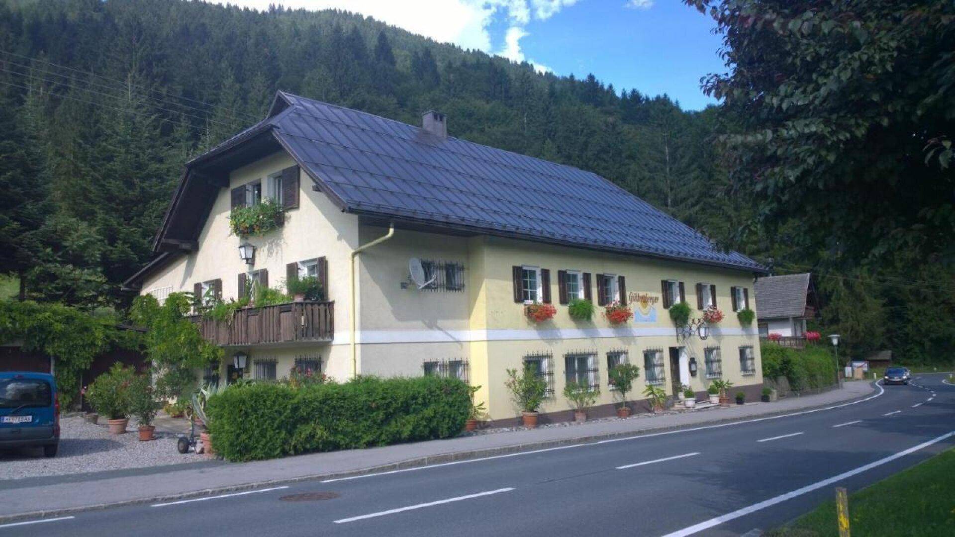 Traditionelles Haus mit gelber Fassade, dunklem Dach und Balkon, umgeben von grünen Bäumen und Bergen.