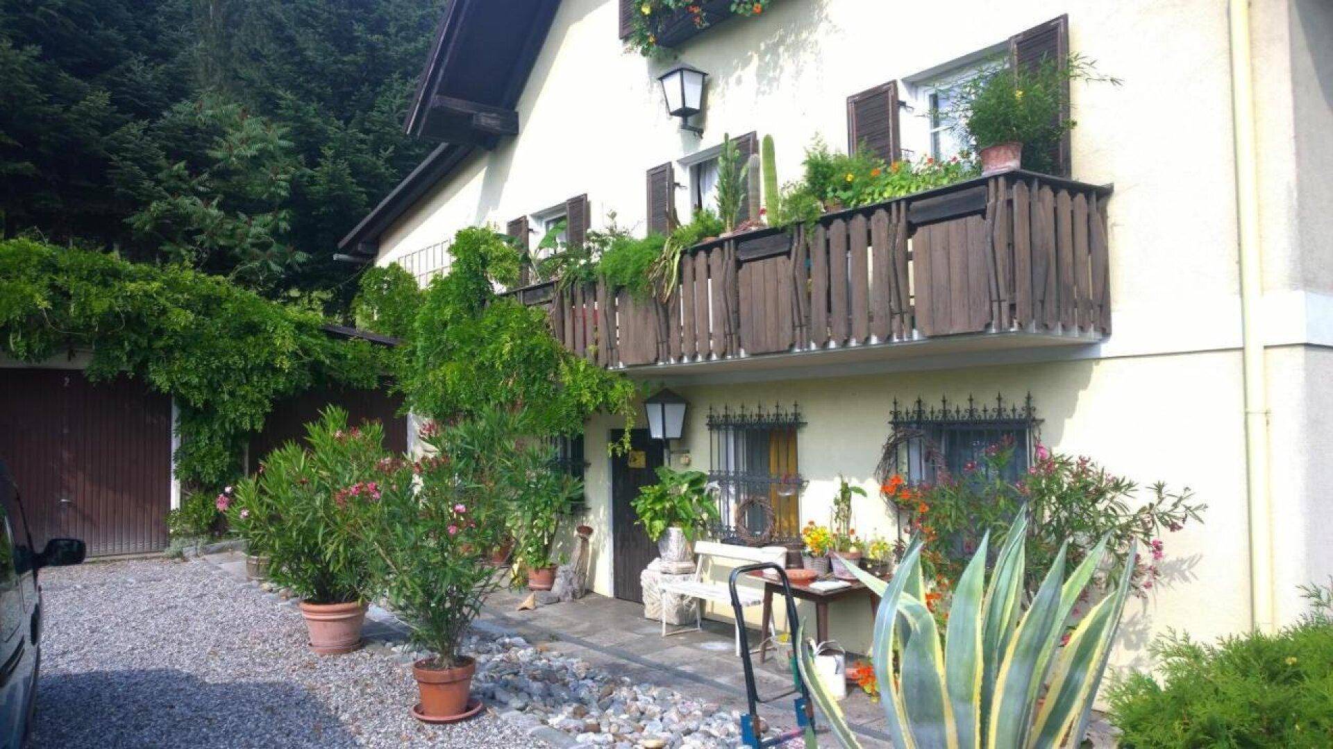 Vorderansicht des Hauses mit Balkon, vielen Topfpflanzen und einem Kiesweg, umgeben von Natur.