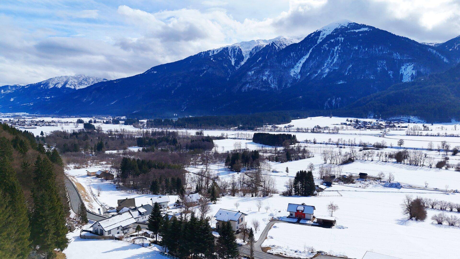 Weitläufige Winterlandschaft mit schneebedeckten Feldern, kleinen Häusern und majestätischen Bergen im Hintergrund.