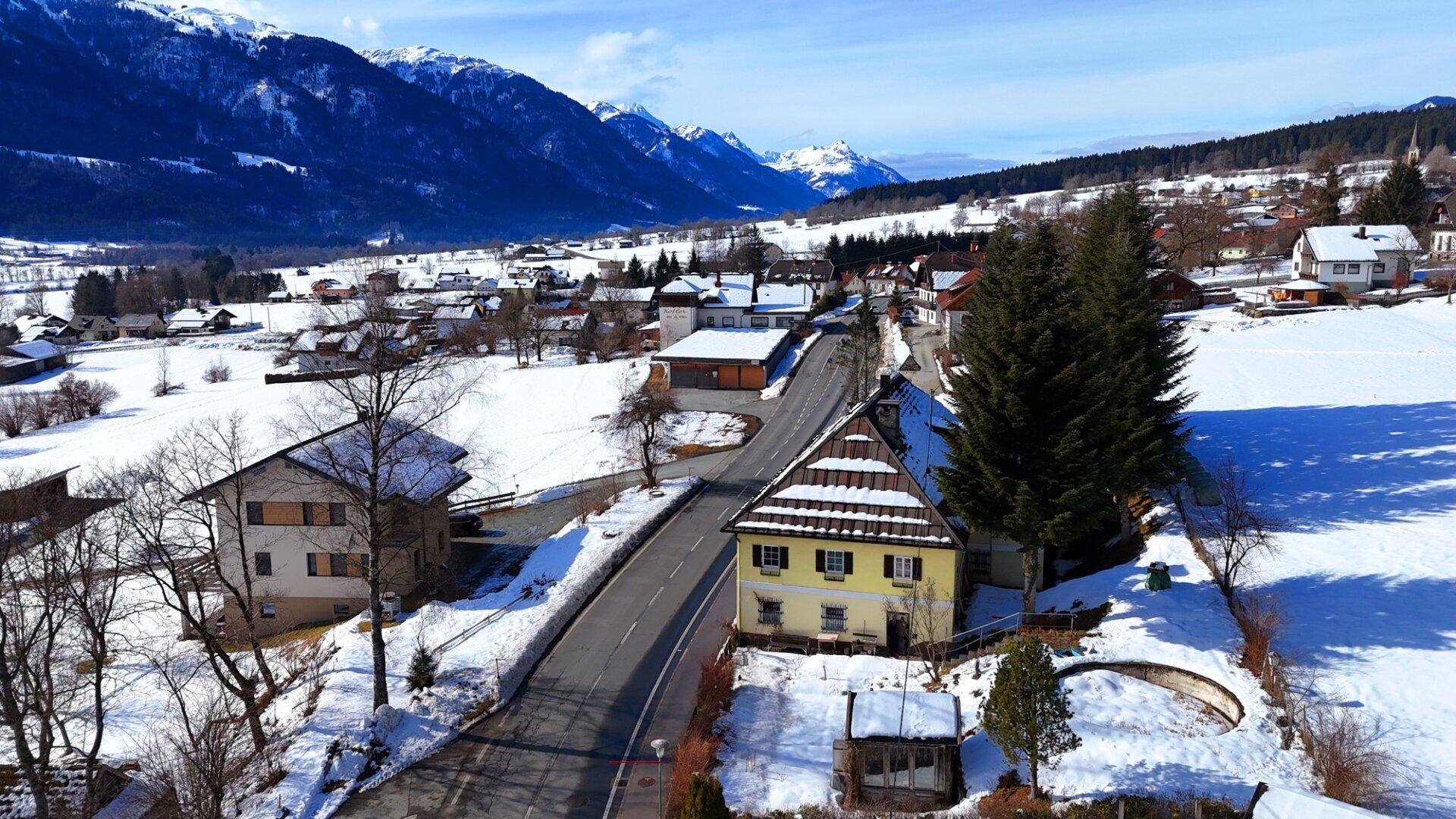 Luftaufnahme eines verschneiten Dorfes mit dem Haus im Vordergrund und einer beeindruckenden Bergkulisse.