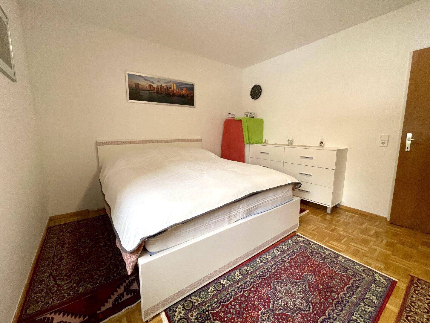 Helles Schlafzimmer mit Doppelbett, Kommode und Holzboden, dekoriert mit Teppichen.