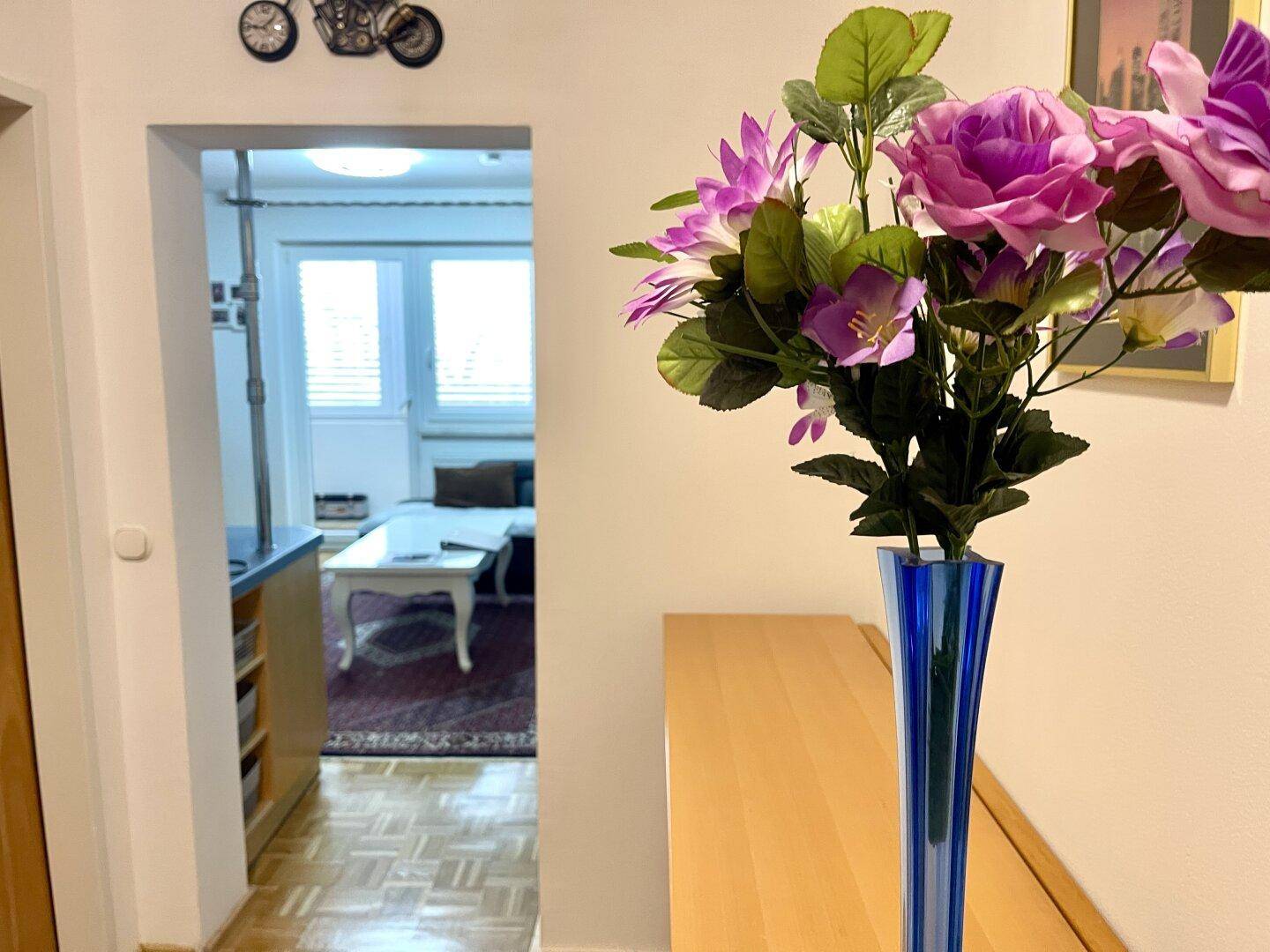 Heller Flur mit Holzboden und einem Blumenstrauß auf einer Holzkommode, der in ein Wohnzimmer führt.