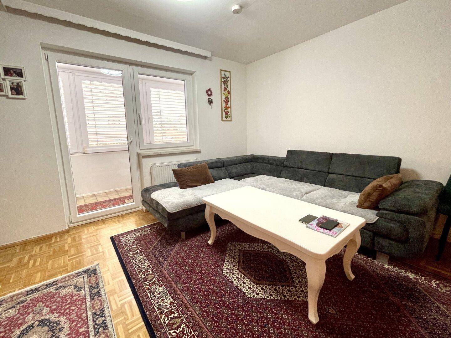 Gemütliches Wohnzimmer mit dunklem Ecksofa, Couchtisch und Zugang zu einem verglasten Balkon.