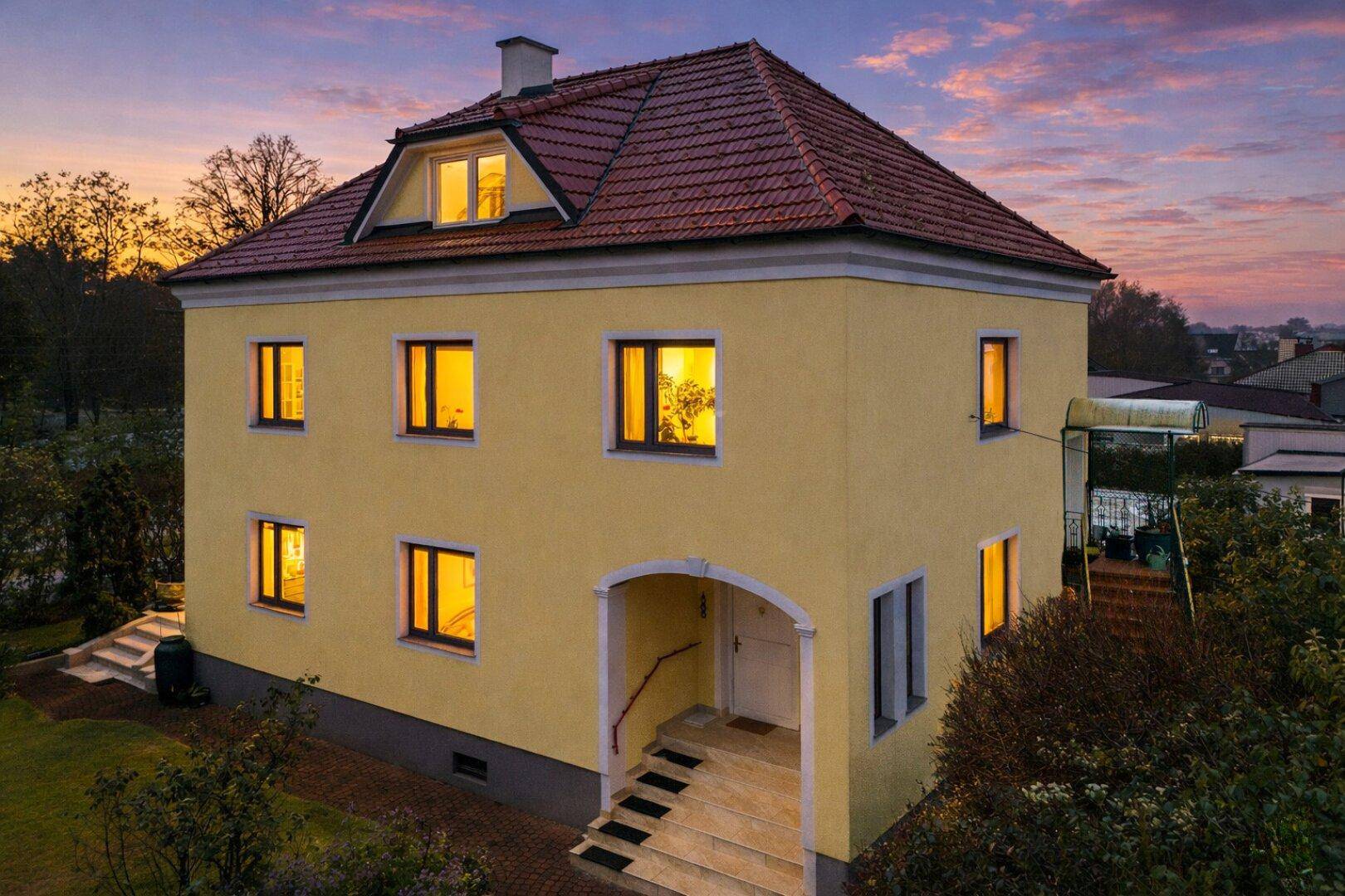 Ansicht der gelben Villa mit beleuchteten Fenstern und einladendem Eingangsbereich bei Sonnenuntergang.