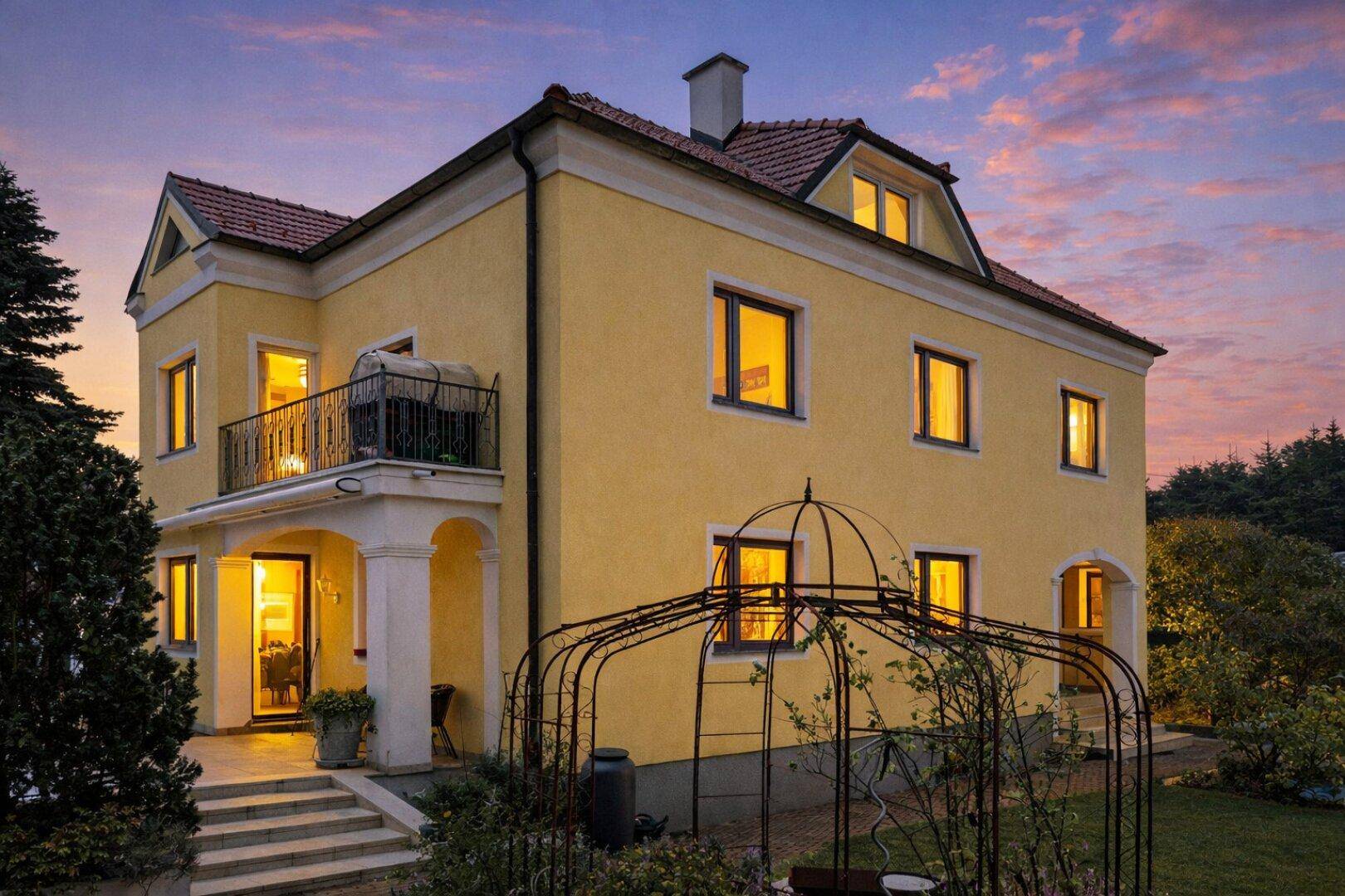 Charmante gelbe Villa mit Balkon und Terrasse im Abendlicht, umgeben von Grünflächen.