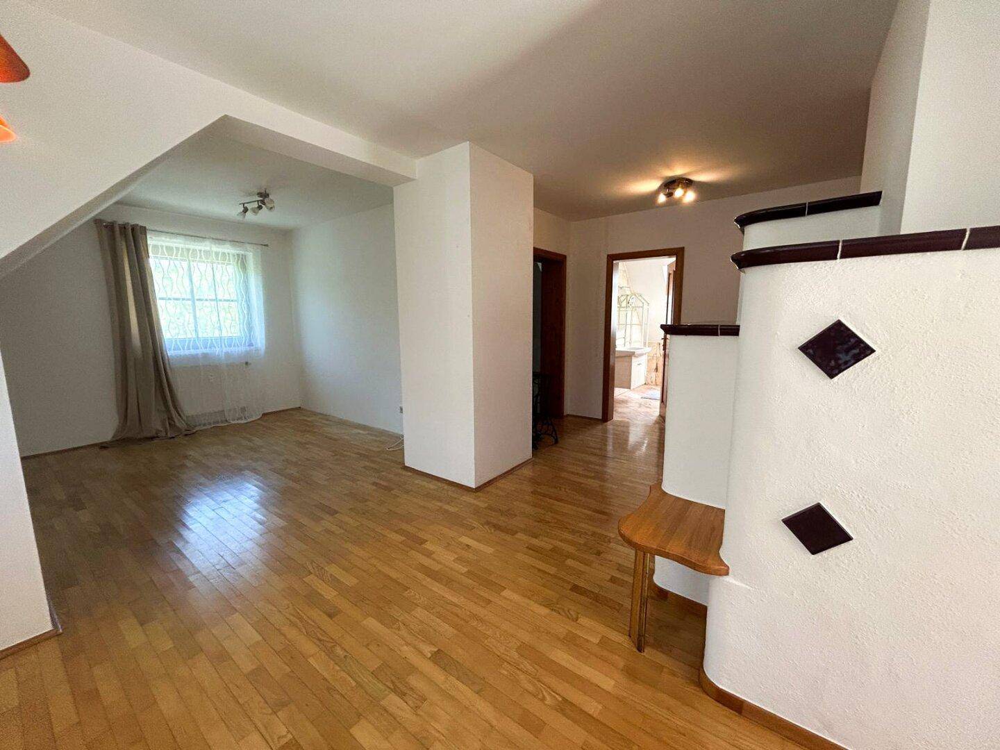 Heller Wohnbereich mit Parkettboden, Fenster und Blick auf den traditionellen Kachelofen.