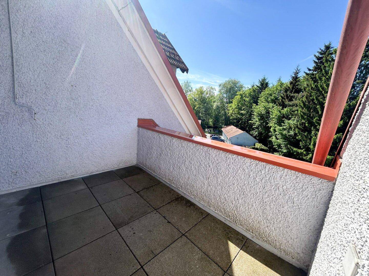 Kleiner Balkon mit Fliesenboden und Blick ins Grüne unter blauem Himmel.