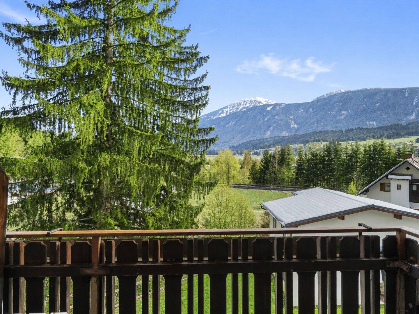 Blick von einem Balkon auf grüne Bäume, Wiesen und schneebedeckte Berge.