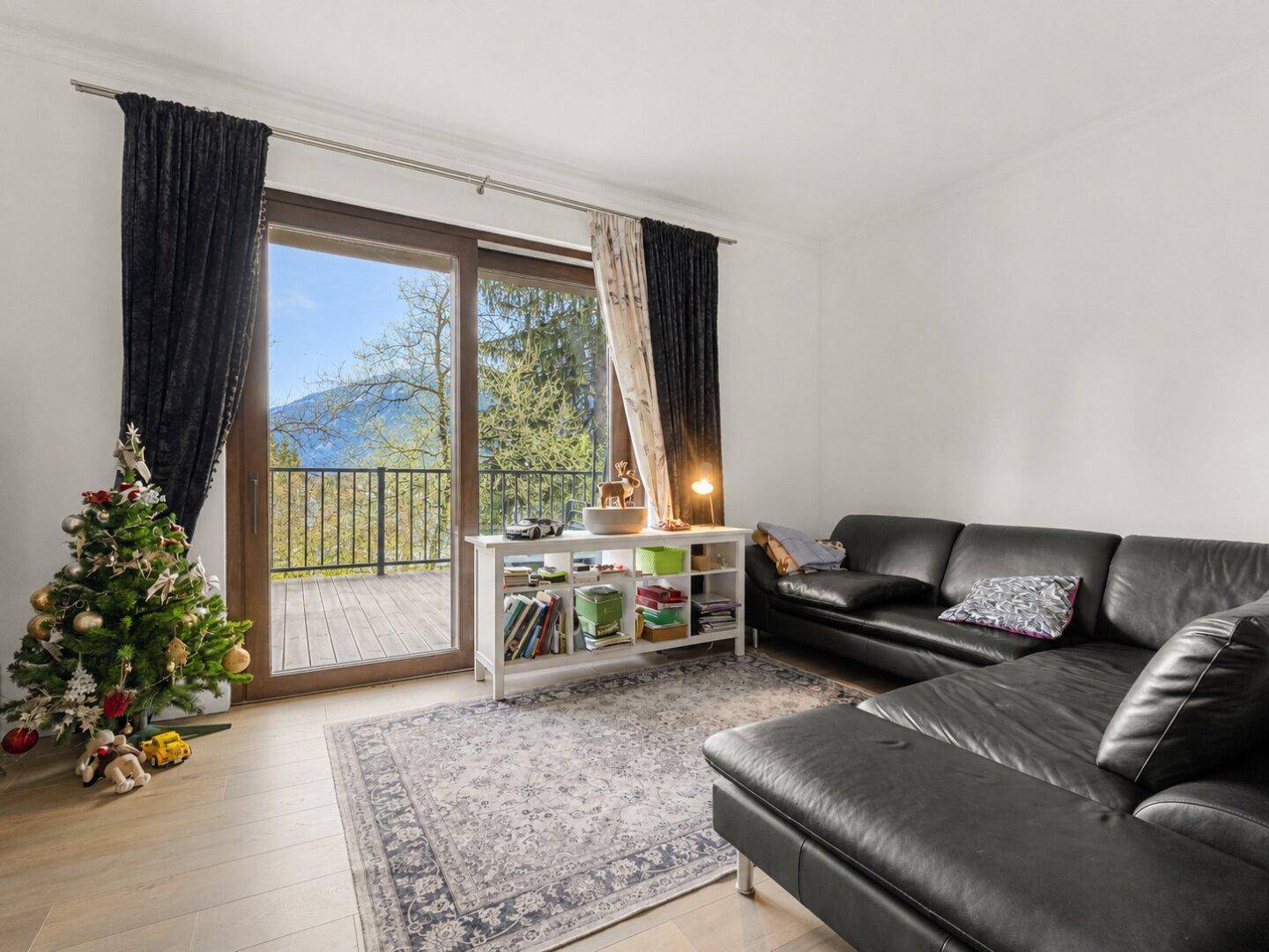 Geräumiges Wohnzimmer mit schwarzem Ledersofa, Teppich und Zugang zu einem Balkon mit Bergblick.