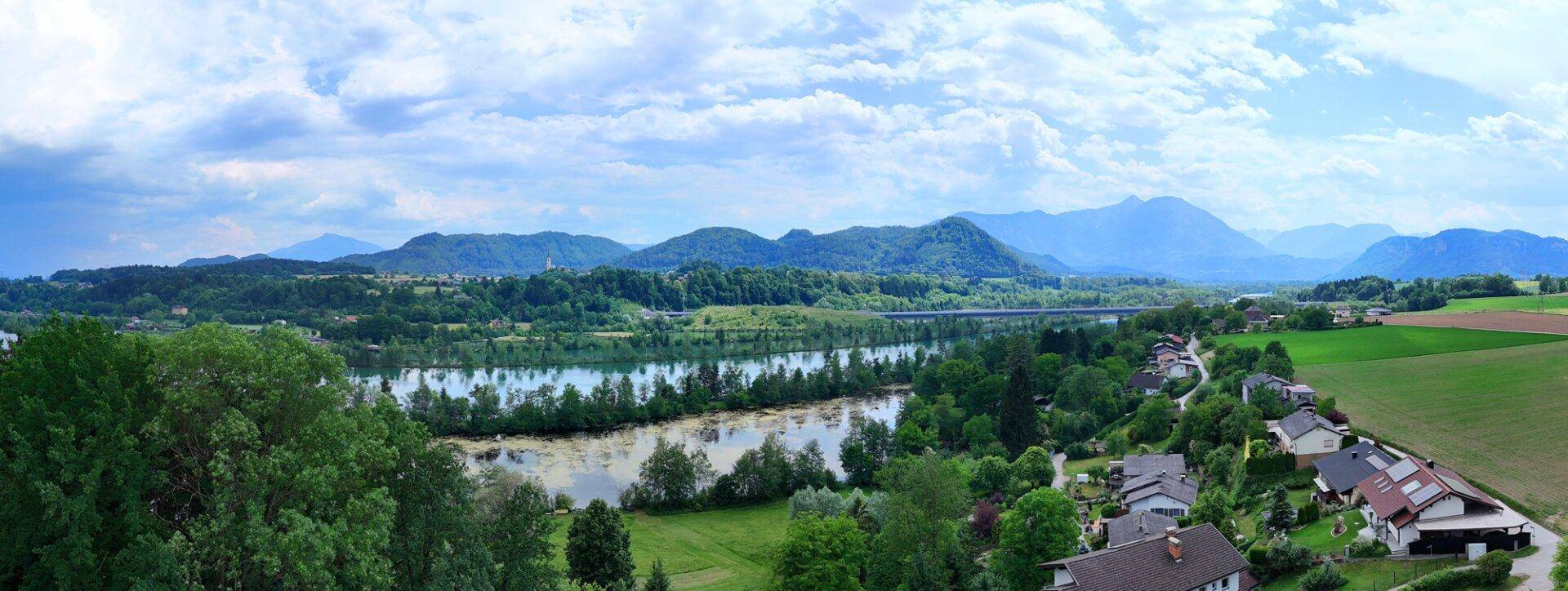 Weitläufiger Panoramablick über einen See, grüne Felder und majestätische Berge im Hintergrund.