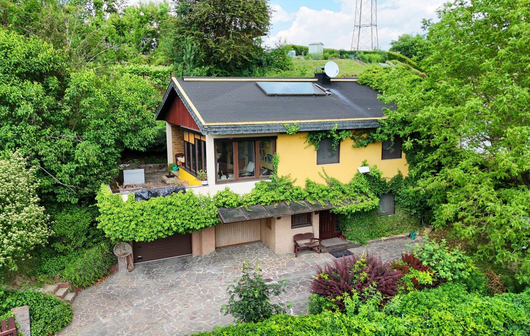 Gelbes Haus mit dunklem Dach, umgeben von Bäumen und einer gepflasterten Zufahrt.